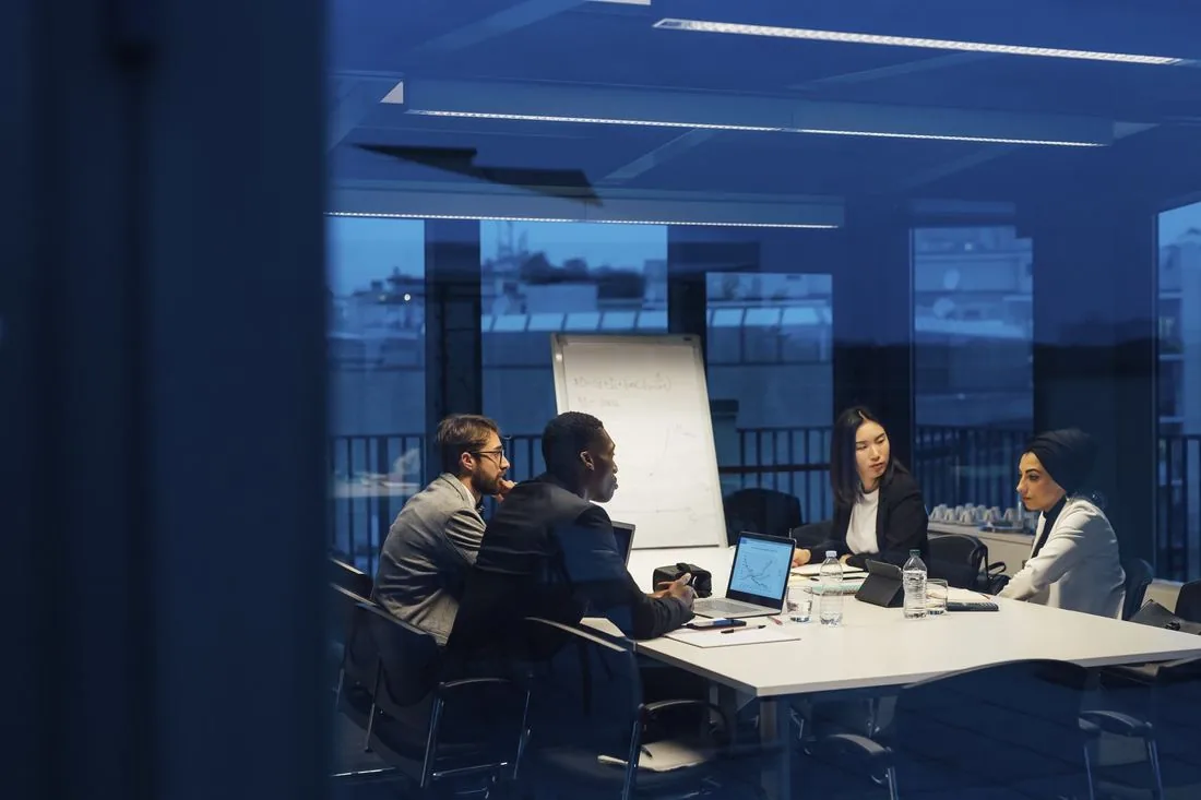 Three people in suits are sitting in a conference room. Their devices are open in front of them and they are discussing their latest efforts in front of a whiteboard.