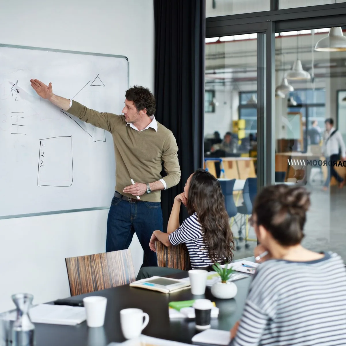 A person stands in front of a whiteboard, presenting ideas to his colleagues in a modern office boardroom. The two women are eagerly listening to his presentation.