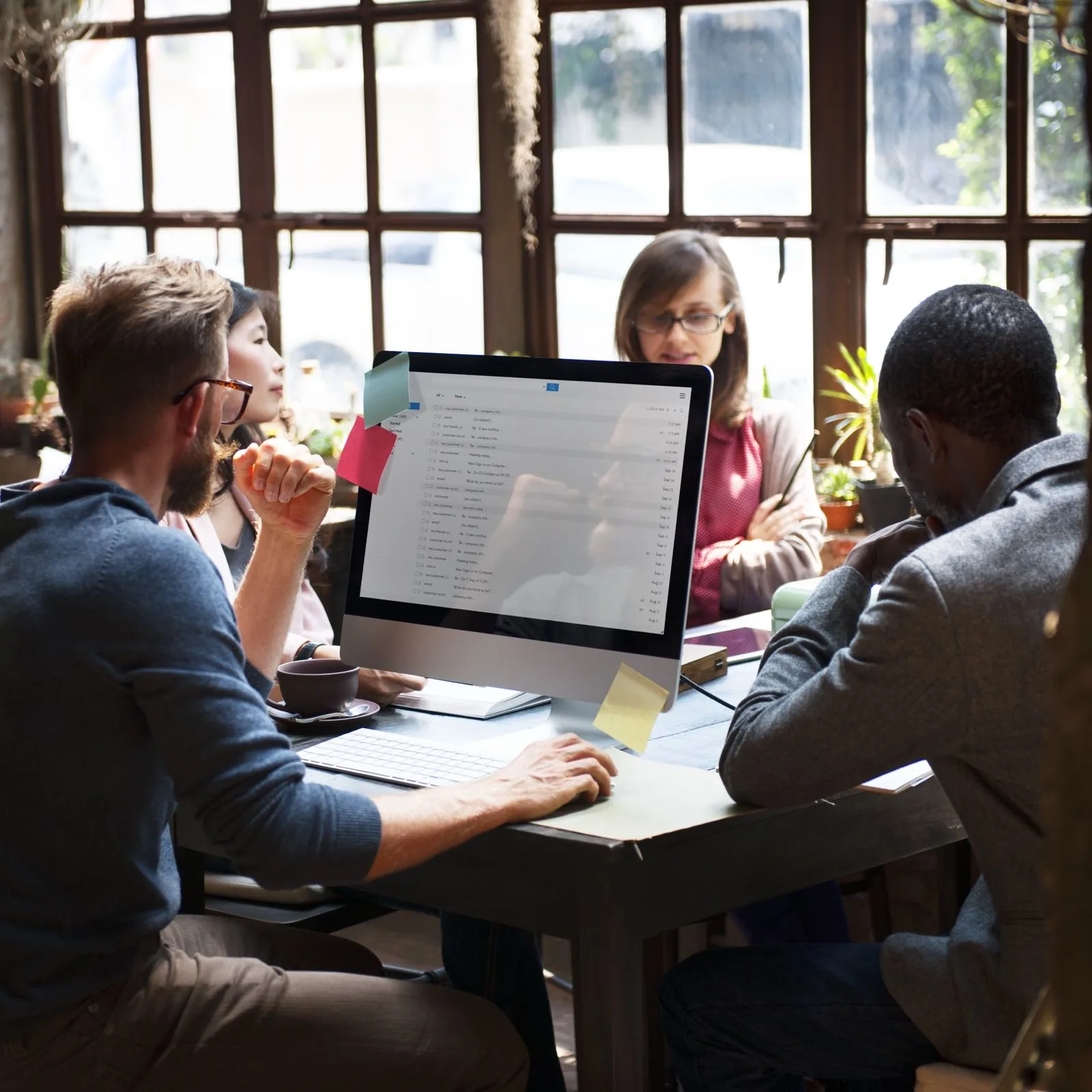 Four people are sitting at a cluttered desk filled with papers and plants. One of them is looking at his computer screen.
