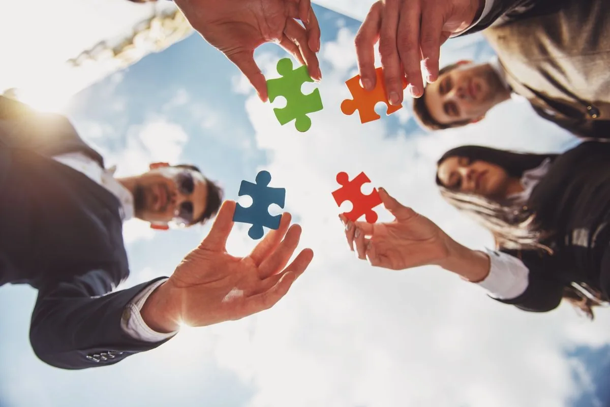 Four people stand around each other, each holding a piece of a puzzle that they&rsquo;re in the process of putting together for team unity.